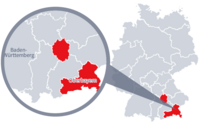 Diese Grafik zeigt eine Karte von Deutschland mit einer detaillierten Ansicht der Region Oberbayern. Oberbayern ist in Rot hervorgehoben und liegt im südlichen Teil Deutschlands. Ein Lupensymbol vergrößert diesen Bereich, um die Lage genauer darzustellen. 